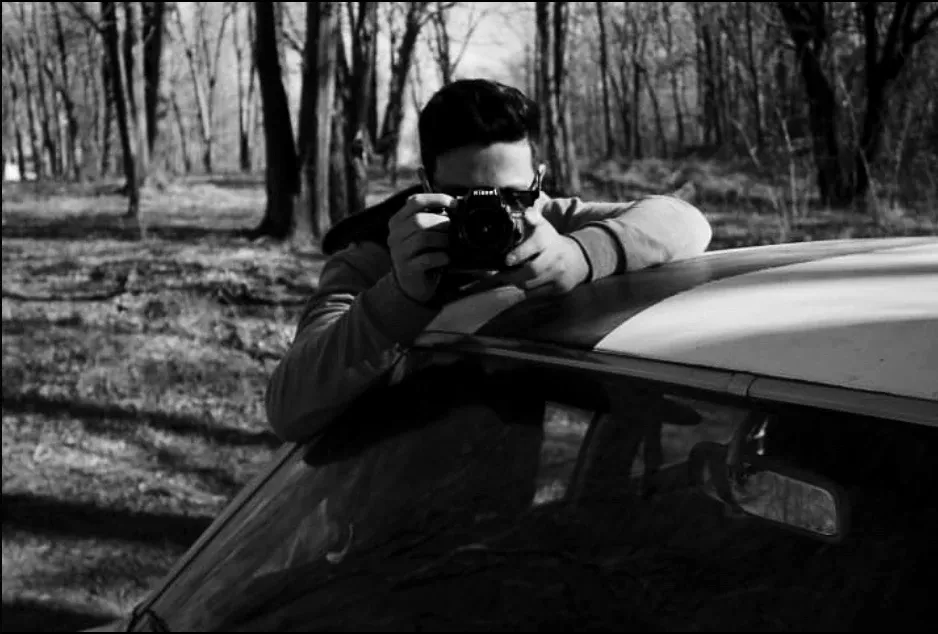 Samu Liendo apoyado en el techo de un auto, apuntando con su cámara Nikon, en blanco y negro