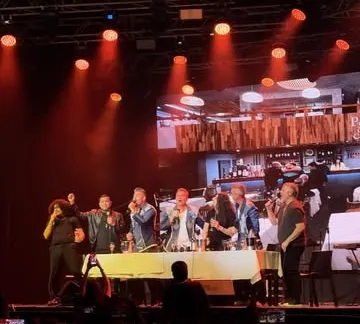 Escenario con luces rojas y los músicos cantando frente a una mesa, con el público filmando con sus celulares
